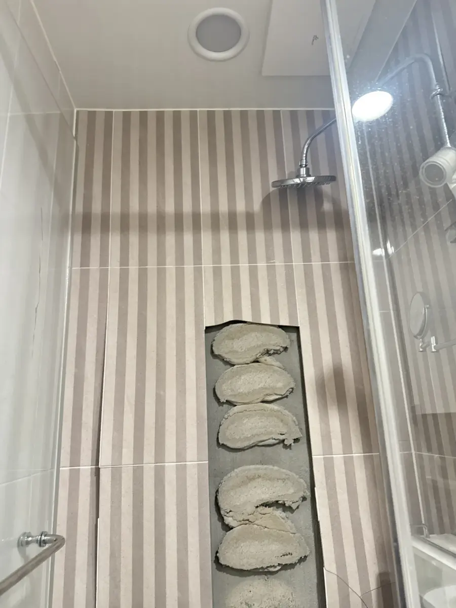 줄무늬 벽타일로 마감된 샤워부스 내부, 하단 니치 선반 자리에 거친 시멘트 마감재 조각이 그대로 노출된 작업 전 실제 현장